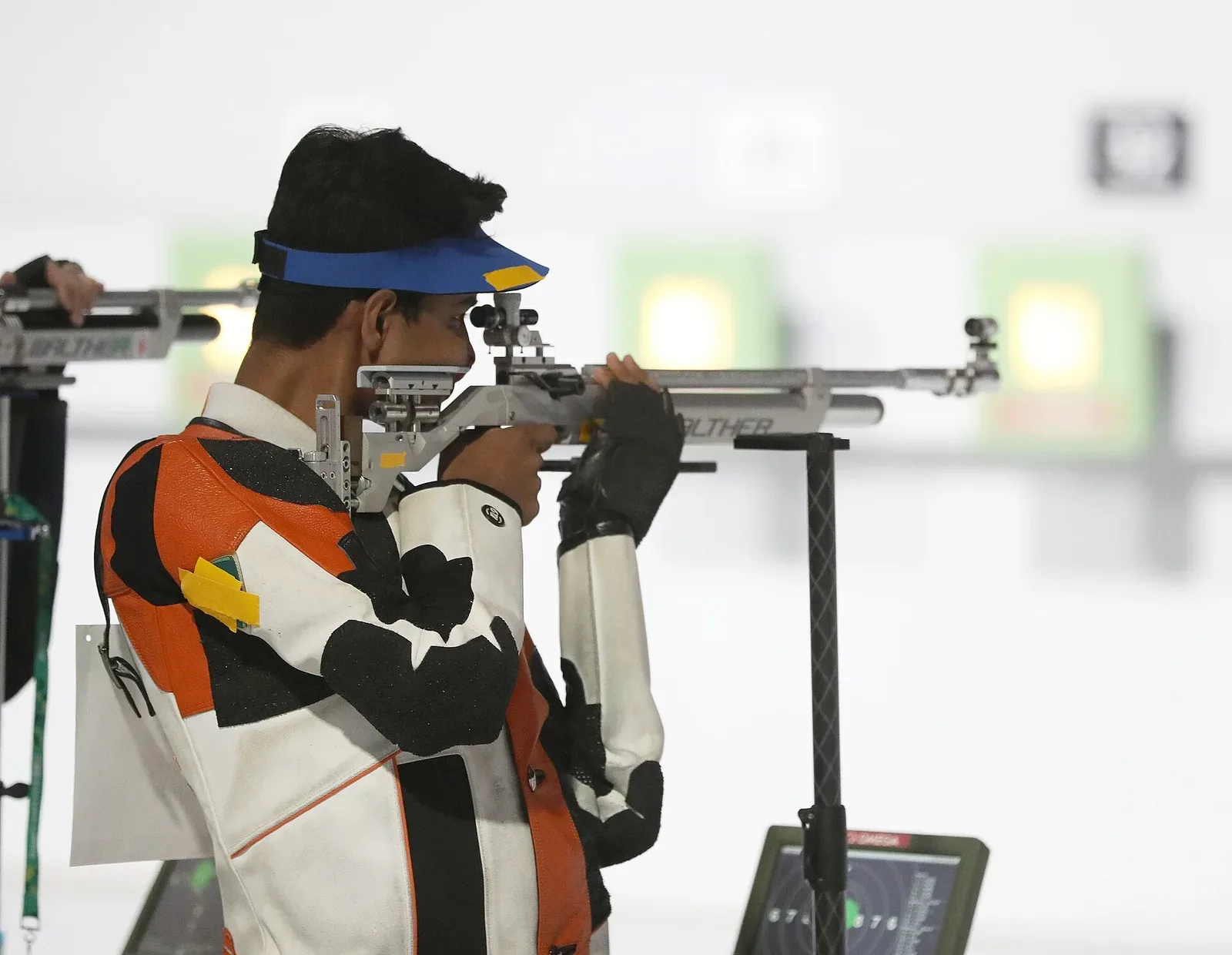 Young male athlete in shooting stance at 10-meter air rifle competition, aiming downrange at an Olympic venue with proper competition setup and positioning