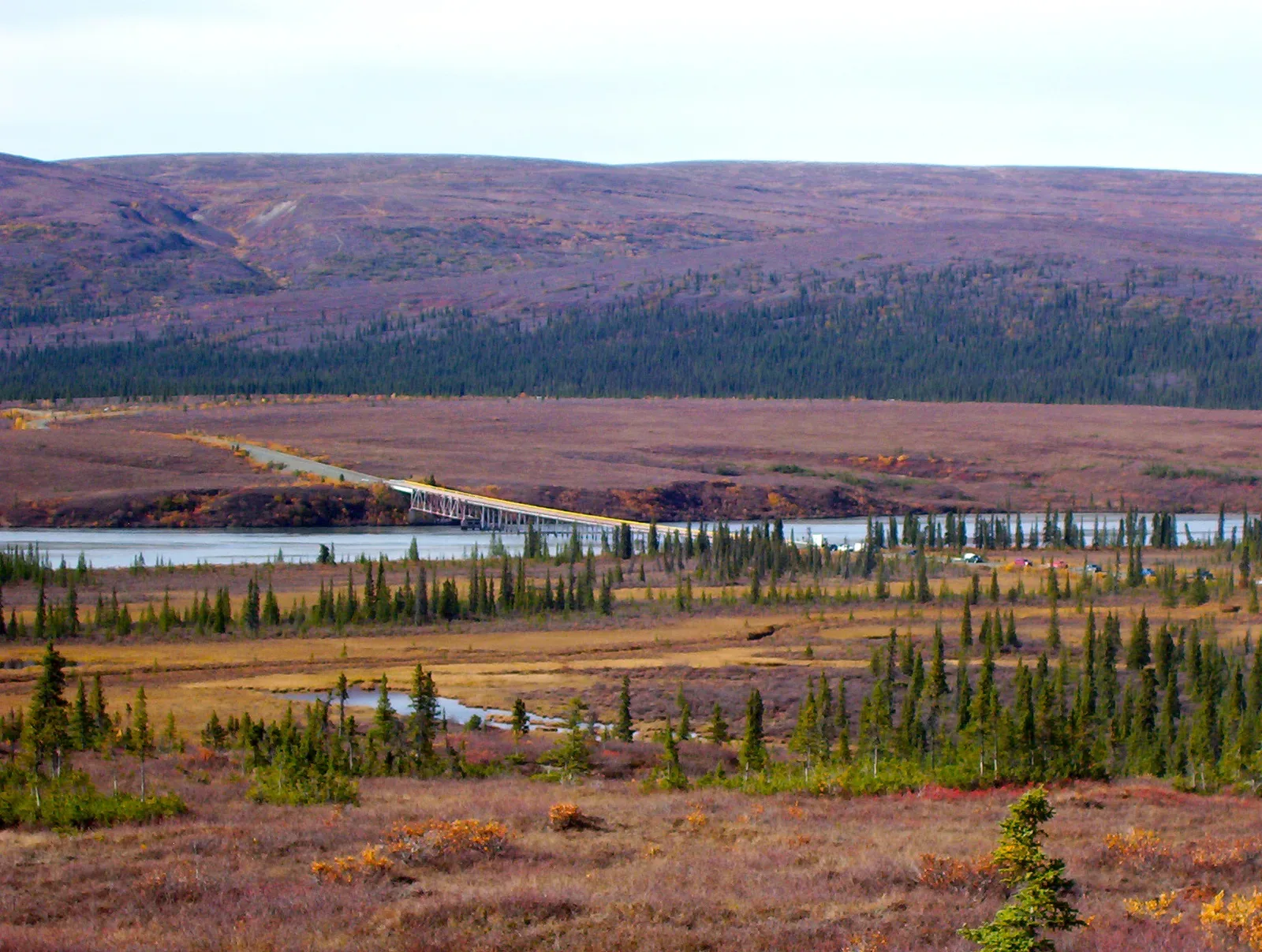 The Denali Highway crossing the Susitna River in Alaska, showing a bridge spanning the river with forested terrain and mountains in the background.