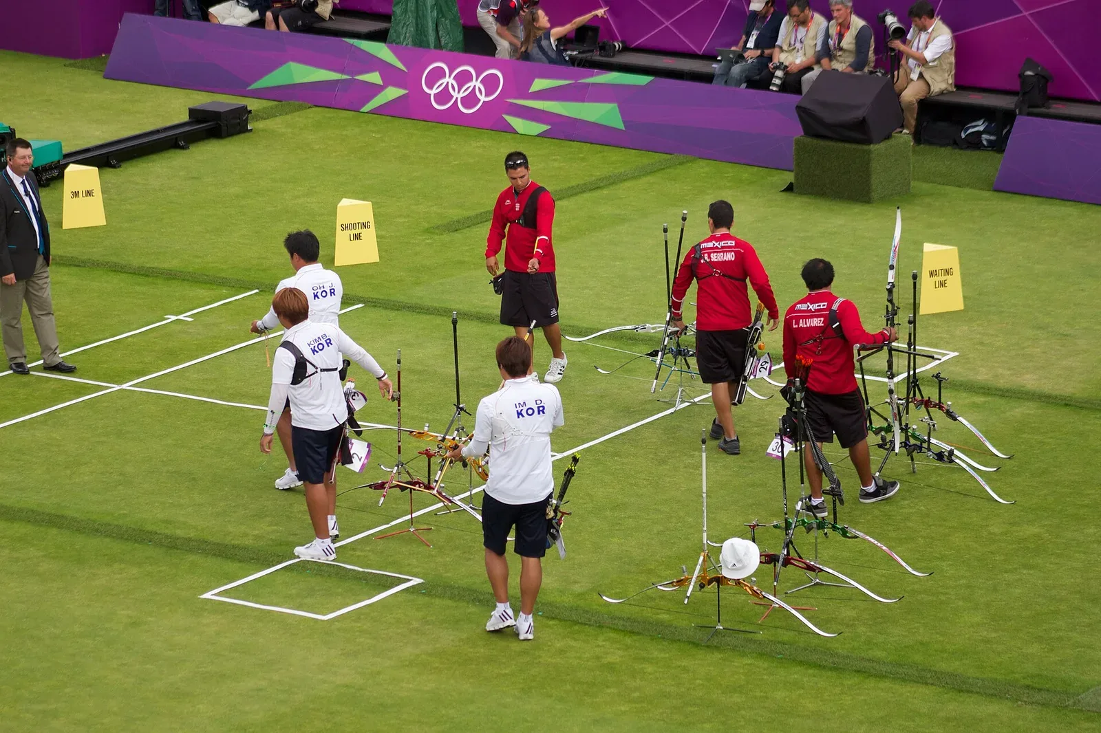 An archer in competition stance drawing a recurve bow during the London 2012 Olympic Games archery event, with spectators visible in the background.