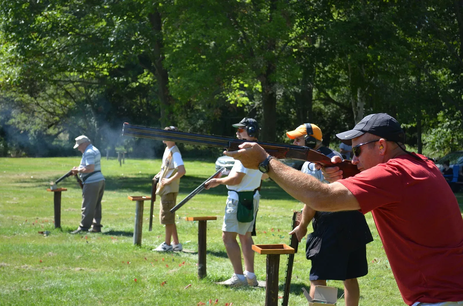 Outdoor trap shooting range with multiple shooters at their stations along the firing line, with trap house visible in the background