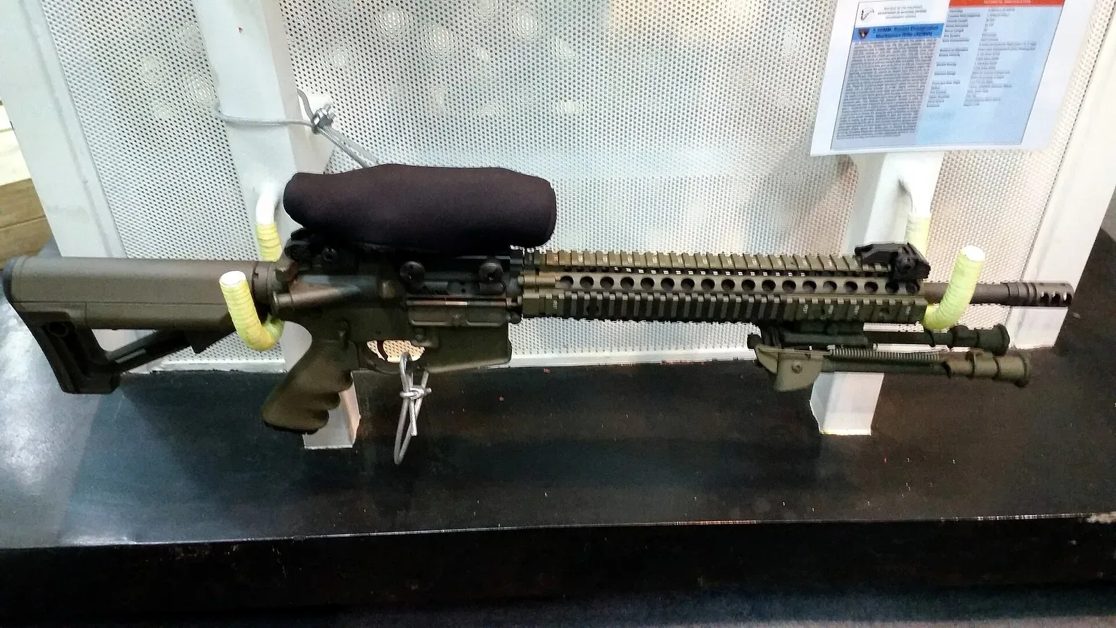 Close-up view of a precision rifle scope mounted on a Squad Designated Marksman Rifle, showing optical lens detail and mounting hardware