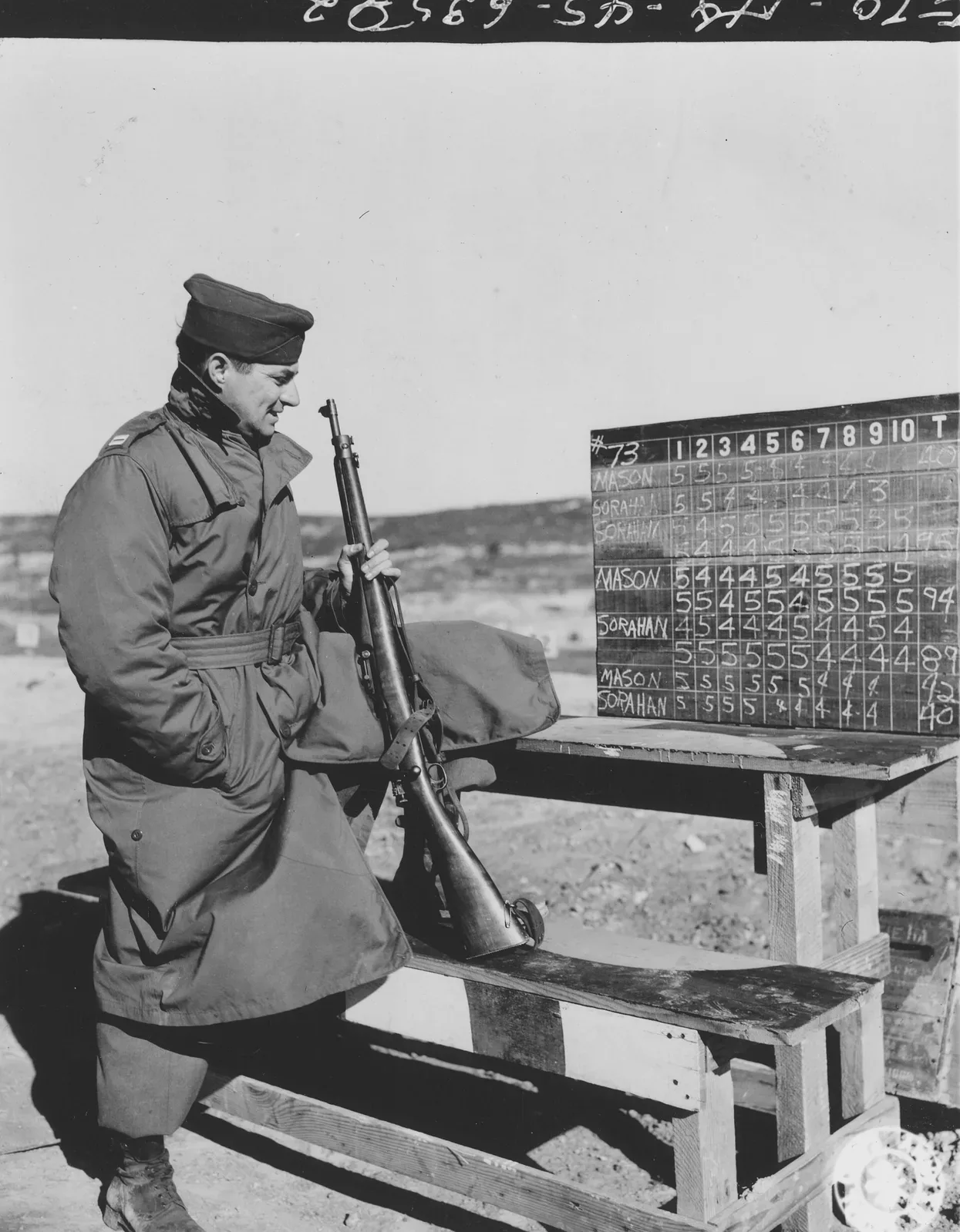 A U.S. Army captain checks a scoreboard while observing rifle competition scores at a shooting range during World War II, with soldiers visible in the background at firing positions.