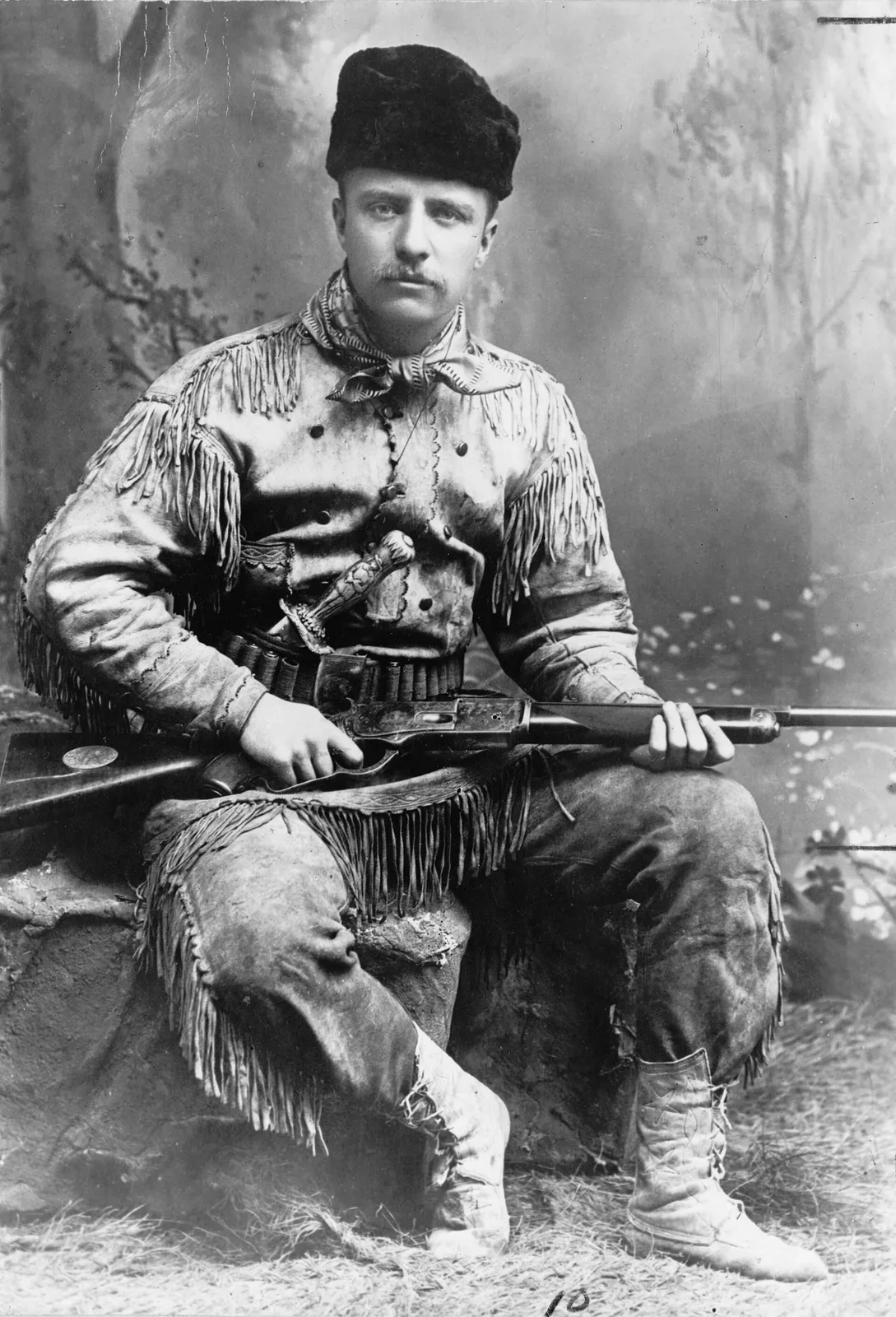 Theodore Roosevelt seated in full-length portrait wearing a deer hunting suit and holding a rifle