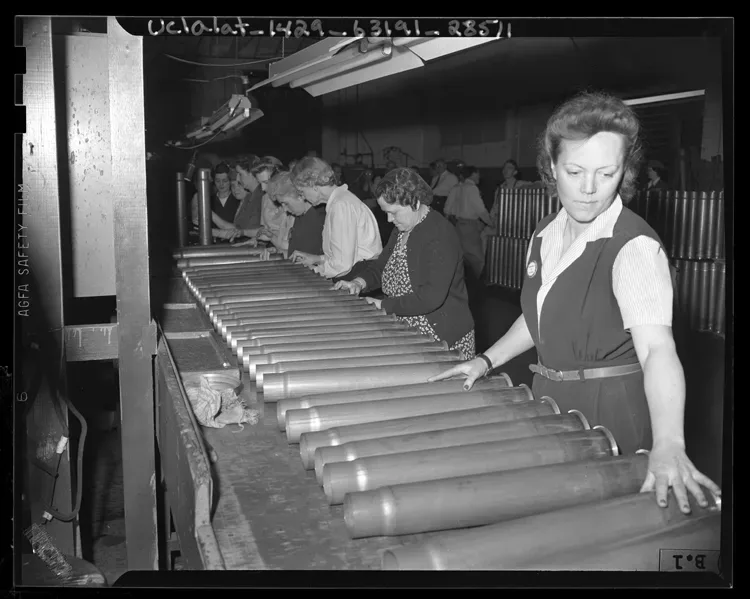 Women ordnance workers inspecting cartridge cases at a manufacturing facility in Los Angeles, California, 1943. Multiple workers are shown examining ammunition components at inspection stations.