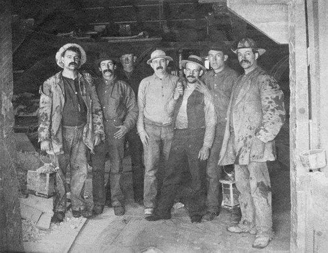 Seven Comstock miners posing for a photograph, circa 1880s, wearing period work clothing and hats typical of late 19th-century mining operations.