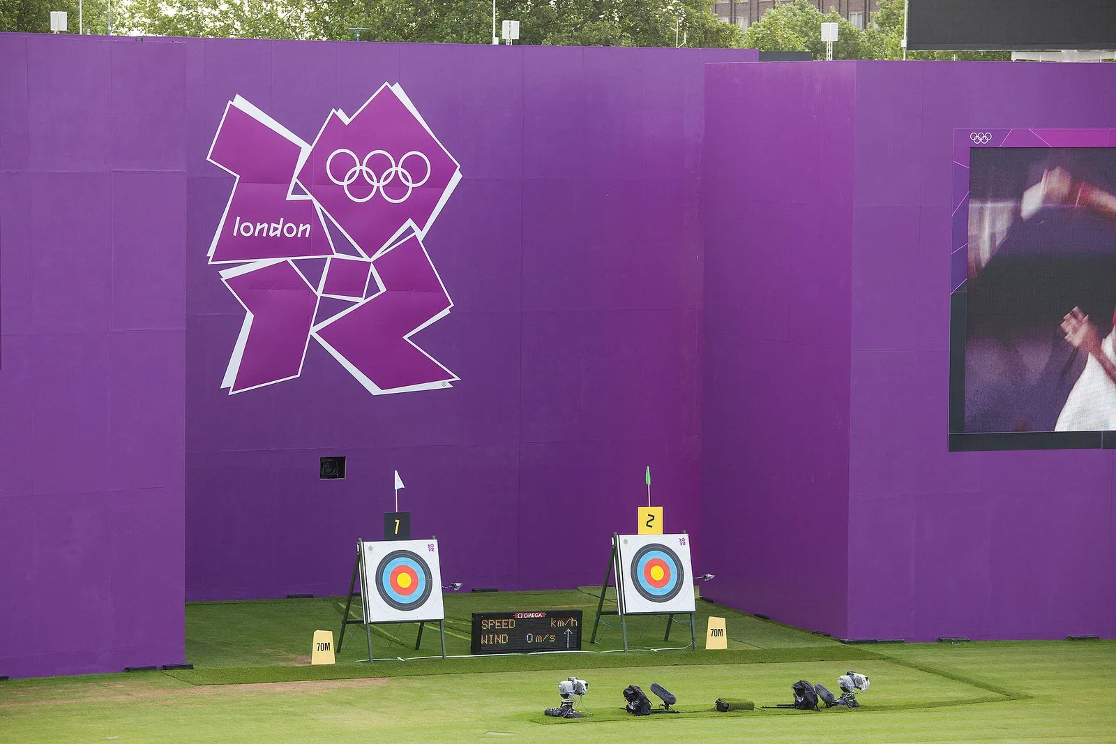 An archer drawing a bow and aiming at a target during competition at the 2012 Summer Olympics, demonstrating proper archery form and technique in an outdoor setting.