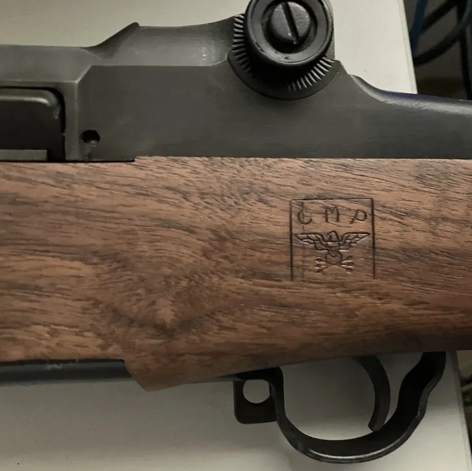 Close-up photograph of a cartouche marking on an M1 Garand rifle showing the Civilian Marksmanship Program insignia stamped into the wood furniture